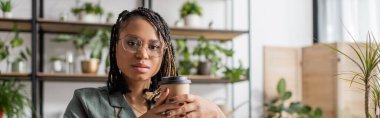 african american florist in stylish eyeglasses and dreadlocks holding coffee to go and looking at camera in flower shop, banner clipart