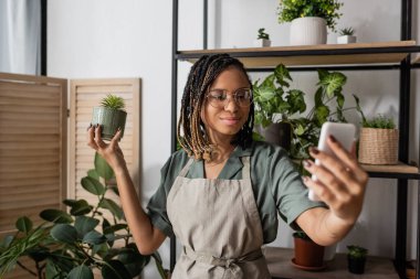 Çiçekçide internetten sipariş verirken elinde saksı saksısı ve cep telefonu tutan şık Afrikalı Amerikalı çiçekçi.