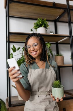 Mutlu Afro-Amerikan çiçekçi elinde saksı bitkisi ve cep telefonu ile çiçekçide videocuda