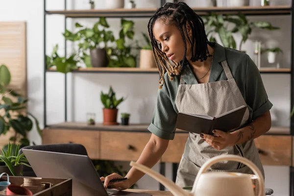 african american florist with trendy hairstyle using laptop and holding copybook near rack with plants on blurred background