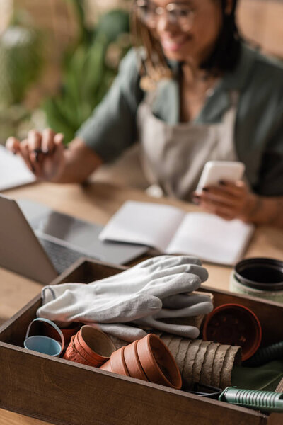 selective focus of box with different flowerpots and work gloves near african american florist with gadgets on blurred background