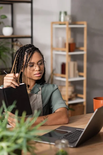 stock image young african american florist in eyeglasses holding notebook and looking at laptop while working in flower shop