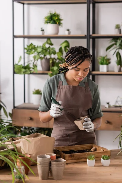african american florist with trendy hairstyle holding garden scoop and flowerpot near wooden box with soil while working in flower shop