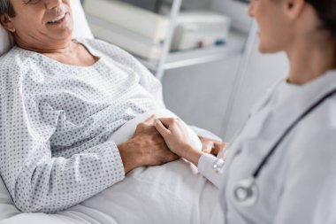 Hastane koğuşunda gülümseyen hastanın elini tutan bulanık bir doktorun görüntüsü. 