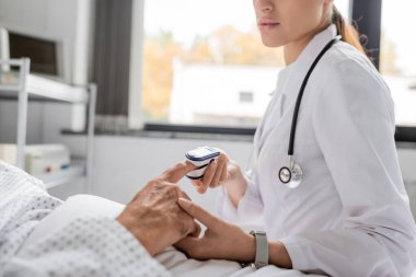 Hastane koğuşundaki yaşlı bir adamın parmağına nabız oksimetresi takan doktor görüşü. 