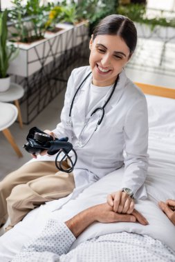 Smiling doctor with tonometer touching hand of senior patient in clinic  clipart