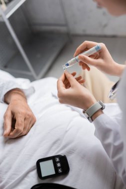 Doktorun elinde tükenmez kalem ve test şeridi tutarken glukometre ve klinikteki hastanın görüntüsü. 