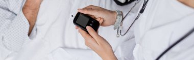 Klinikte yaşlı bir hastanın yanında glukometre tutan doktor görüntüsü. 
