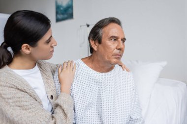 Woman hugging upset dad in patient gown in clinic  clipart