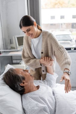 Positive woman holding hand of elderly father in patient gown in clinic  clipart