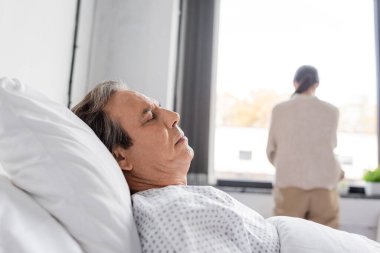 Senior man lying on bed near blurred daughter in hospital ward  clipart