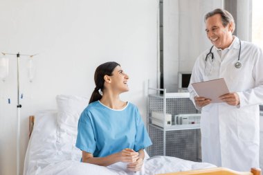 Smiling senior doctor holding paper folder near patient in hospital ward  clipart