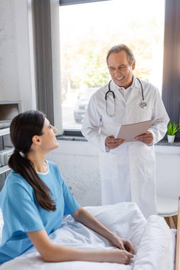 Elinde kağıt klasörü olan yaşlı doktor hastane koğuşundaki hastalarla konuşuyor. 
