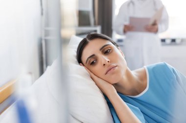 Dissatisfied patient lying on bed near blurred doctor in hospital ward  clipart