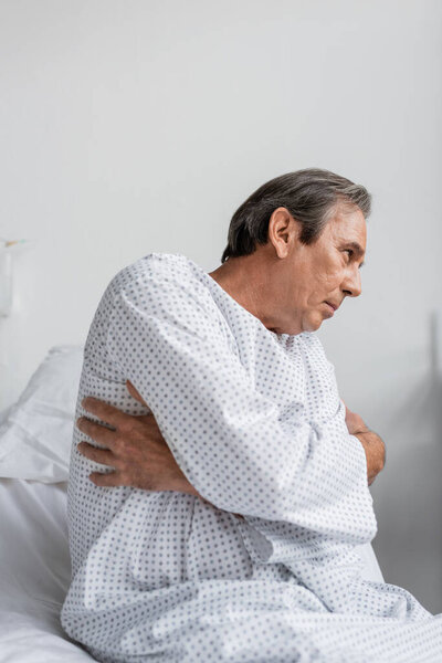 Side view of sick senior man sitting on bed in hospital ward
