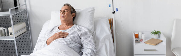 Elderly man in patient gown lying near intravenous therapy and pills in clinic, banner