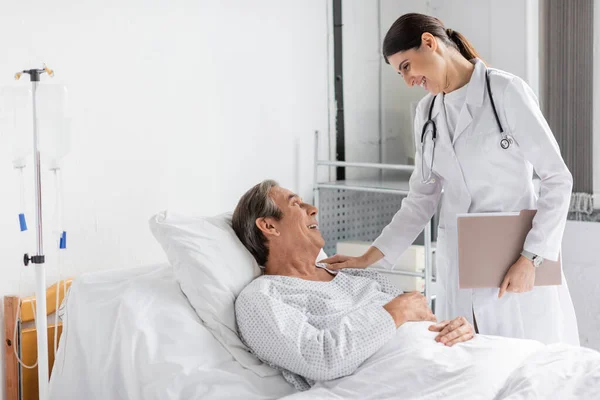 Side view of smiling doctor with paper folder calming elderly patient in hospital ward