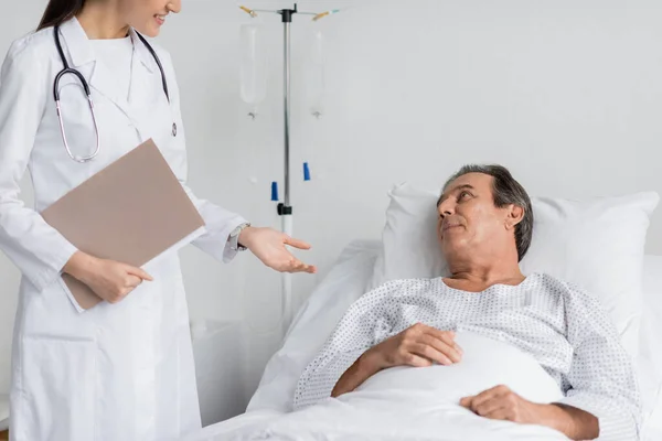 Smiling doctor with paper folder talking to senior patient on bed in clinic