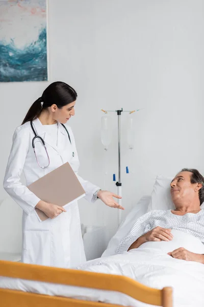brunette doctor with paper folder talking to elderly patient in clinic