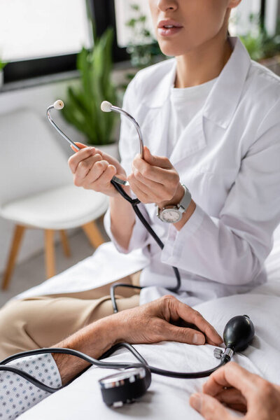 Cropped view of doctor holding stethoscope near senior patient and tonometer on hospital bed 