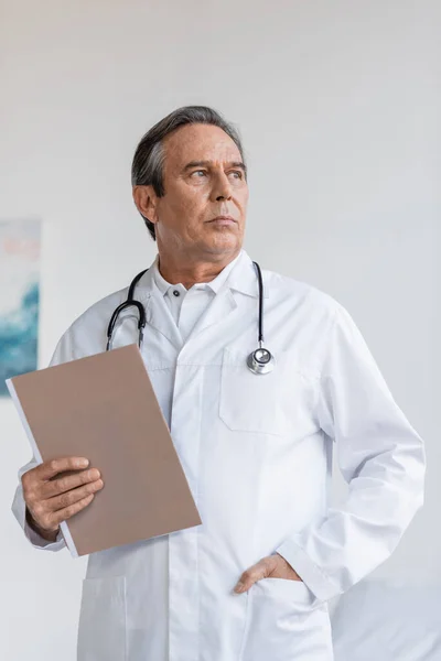 Senior doctor holding paper folder and looking away in clinic