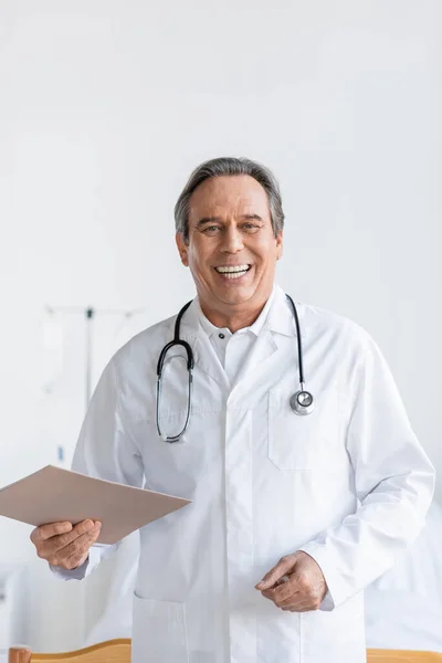 Positive senior doctor holding paper folder and looking at camera in clinic
