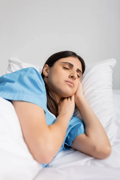 Ill brunette patient in gown sleeping while lying on bed in clinic
