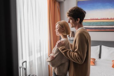 side view of smiling man hugging girlfriend standing with crossed arms near window in hotel room