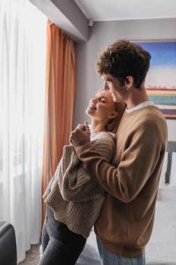 happy blonde woman with closed eyes holding hands with smiling boyfriend in hotel apartments
