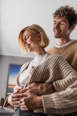 low angle view of smiling man hugging cheerful blonde girlfriend in hotel apartments