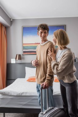 smiling man looking at blonde girlfriend standing with suitcase in hotel bedroom