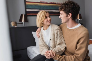 young travelers holding hands and smiling at each other while sitting in hotel bedroom