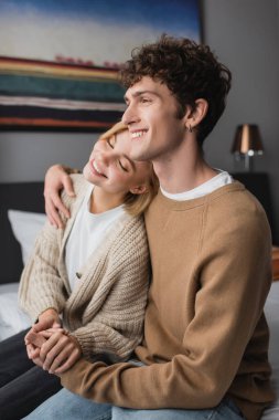 smiling man holding hands with happy girlfriend sitting with closed eyes on bed in hotel