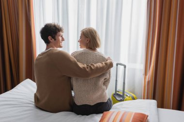 back view of cheerful man hugging blonde girlfriend while sitting on bed in hotel apartments
