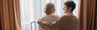 back view of brunette man hugging happy blonde girlfriend in hotel apartments, banner