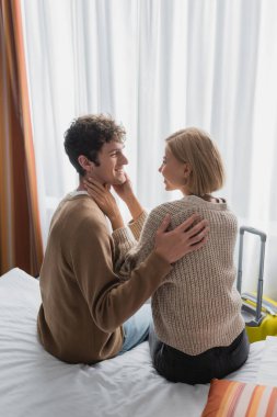 pleased young couple sitting on bed in hotel apartments and smiling at each other