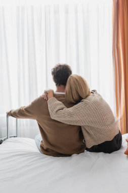 back view of blonde woman hugging boyfriend sitting on bed in hotel near suitcase