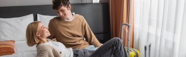 young blonde woman smiling on bed near boyfriend in hotel apartments, banner