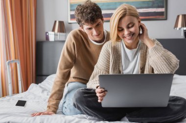 smiling blonde woman sitting with laptop on hotel bed near young boyfriend 