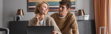 blonde woman and brunette man smiling at each other near laptop in hotel apartments, banner