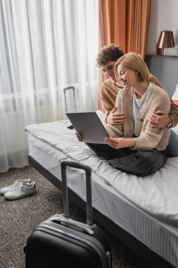 young man hugging girlfriend sitting with crossed legs and laptop on bed in hotel apartments