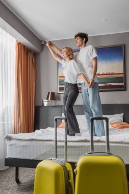 full length of joyful couple standing on bed and holding hands while having fun in hotel room near travel bags