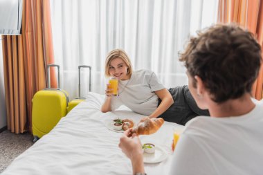 smiling woman looking at blurred boyfriend while having breakfast with orange juice and croissant on bed in hotel