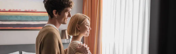 stock image side view of happy man hugging shoulders of smiling blonde girlfriend near window in hotel suite, banner