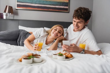 young man using mobile phone near smiling girlfriend and breakfast on bed in hotel