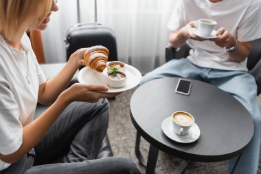 cropped view of smiling woman holding croissant with jam and chocolate paste while having breakfast near blurred man in hotel clipart