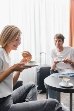 cheerful woman holding tasty croissant near boyfriend with coffee cup in hotel room on blurred background