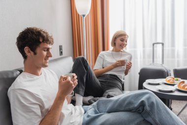 young couple with coffee cups sitting on couch near tasty croissants in hotel room