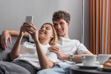 cheerful blonde woman using cellphone near young boyfriend and blurred coffee cups on couch in hotel