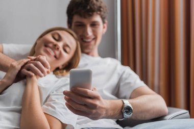 young and happy man holding cellphone while resting and holding hands with girlfriend on blurred background in hotel room